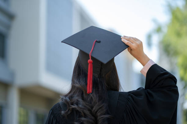A person wearing a graduation cap and gown is shown from behind. They are holding the cap in one hand, and the cap features a red tassel. The background displays blurred urban buildings with greenery, symbolizing the celebration of academic achievement.